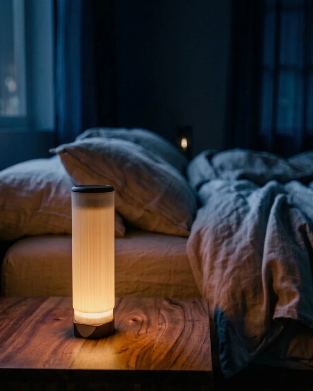 ambielo+ glowing on a walnut nightstand beside an unmade linen bed at night